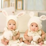 Twin babies wearing cute bunny outfits with plush toys indoors.