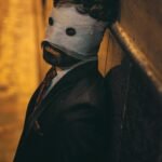 A suited man with a bandaged face stands against a dimly lit wall, evoking mystery.