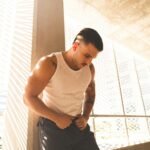 A stylish young man with tattoos poses in bright sunlight indoors.