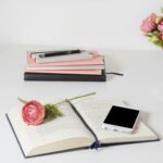 A stylish home office setup with open book, smartphone, and flowers on a white desk.