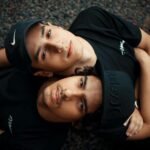 Two young men in casual attire, lying on gravel, showcasing friendship and style.