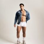 A fashionable male model wearing a denim jacket and white underwear, posing confidently.