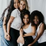 Four diverse women embrace, smiling, showcasing friendship and unity indoors.