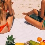 Two women in bikinis enjoying a sunny beach day with fruits and a book.